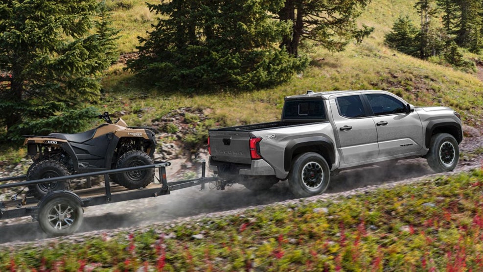A silver Toyota Tacoma towing a trailer with a dirt bike up a road on a hill.