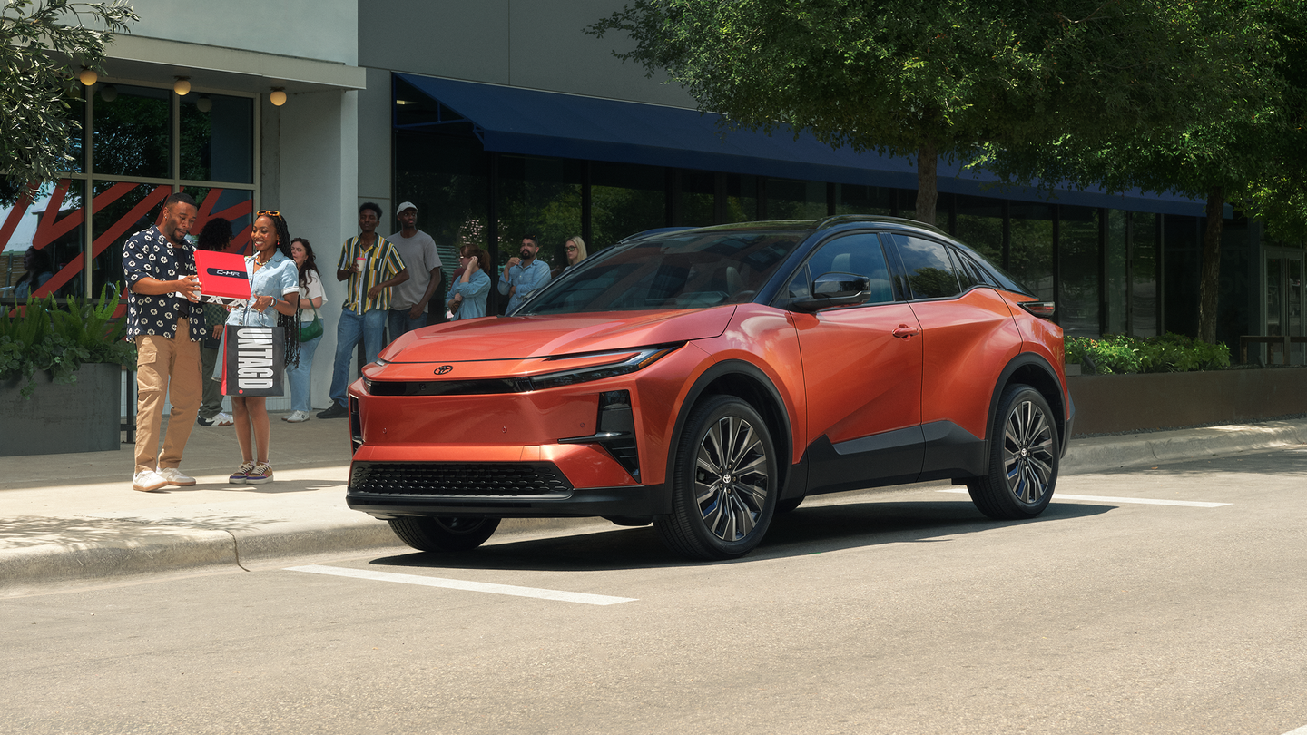 A red-orange Toyota CH-R parked by the curb near a group of people.