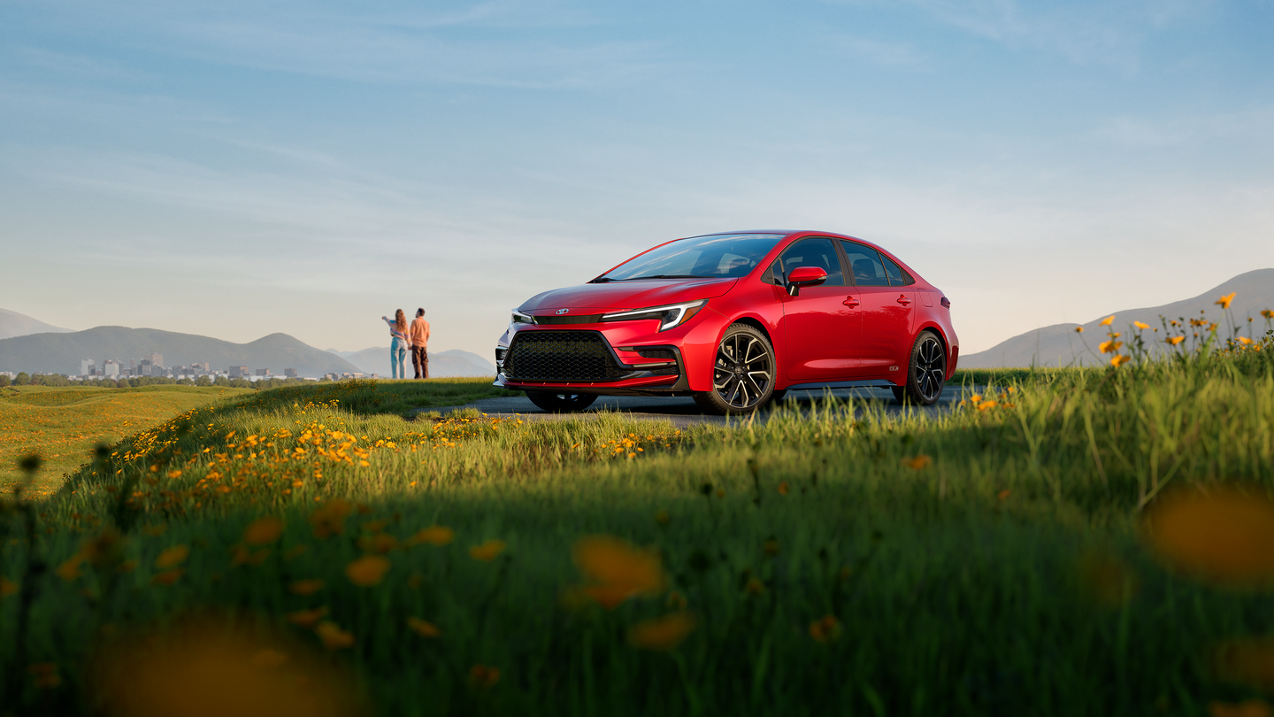 A red Toyota Corolla parked in a field with a couple looking at a city in the distance.