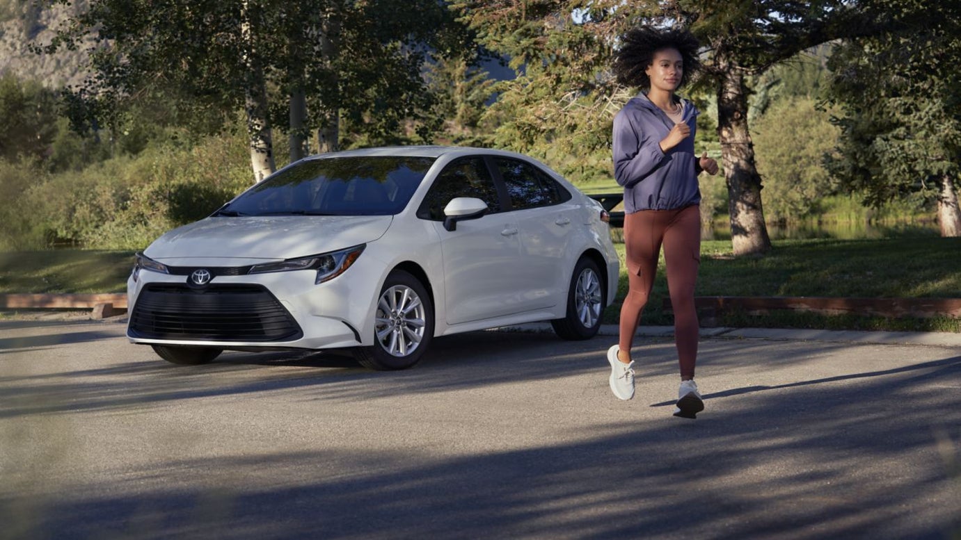 A white Toyota Corolla parked by a park while a woman jogs away from it