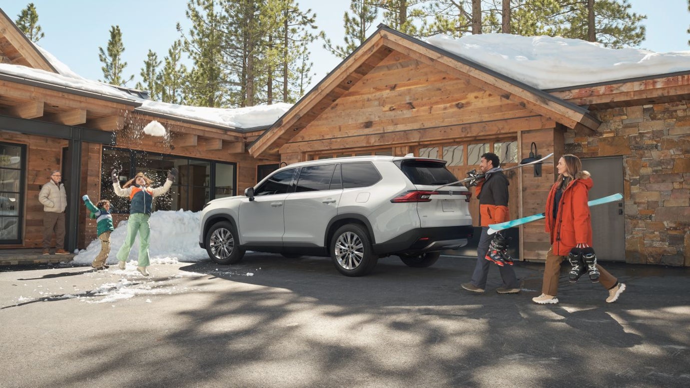 A silver Toyota Grand Highlander in front of a snow-covered cabin near a family.