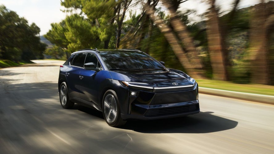 A blue Toyota bZ driving down the road by a forest.
